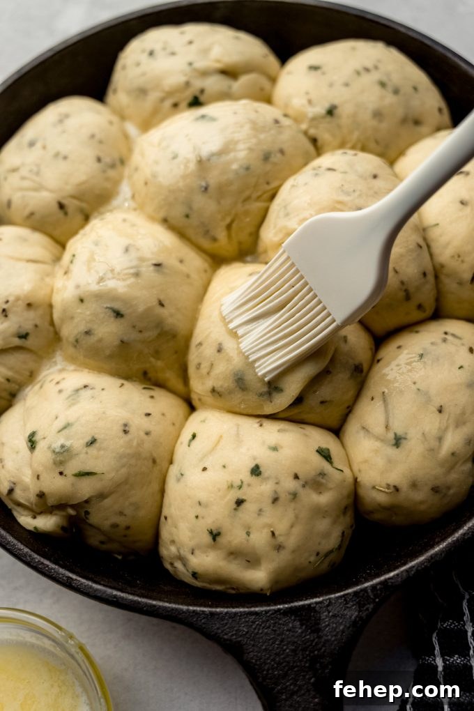 Someone is brushing melted butter onto balls of herbed roll dough.