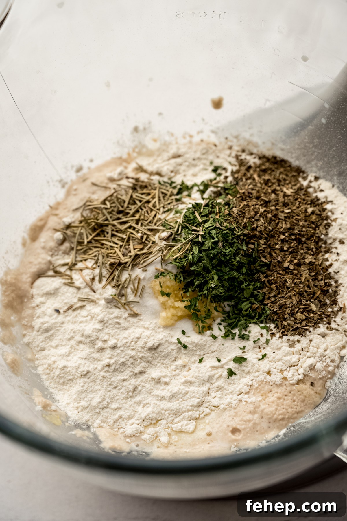 Herbs and flour and other ingredients in a bowl of a stand mixer making herbed rolls.