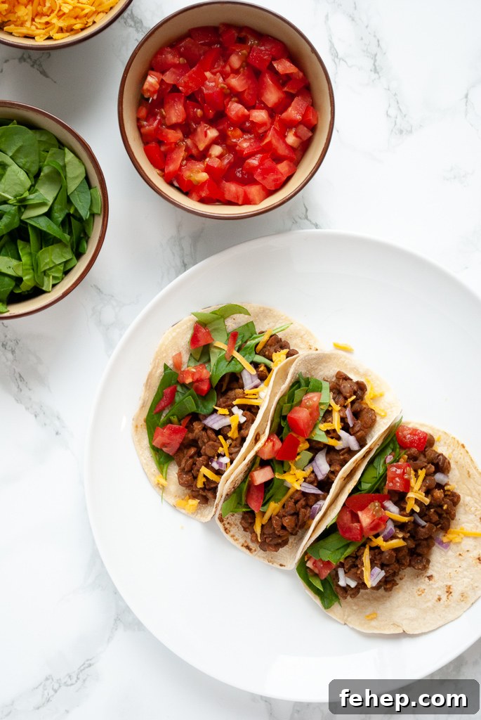 A top down view photo of a plate of lentil tacos with bowls of chopped tomato and spinach to the side of it.