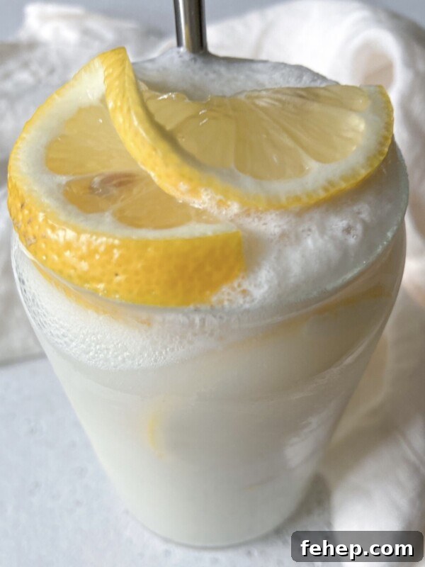 A creamy, frothy lemon drink served in a clear glass, garnished with two fresh lemon slices on top, with a white cloth in the background.