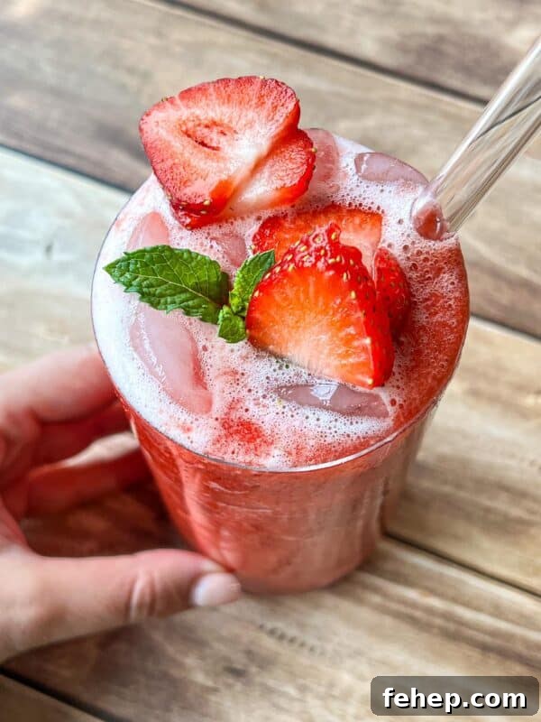 A hand holds a glass of iced strawberry drink topped with fresh strawberry slices and a mint sprig.