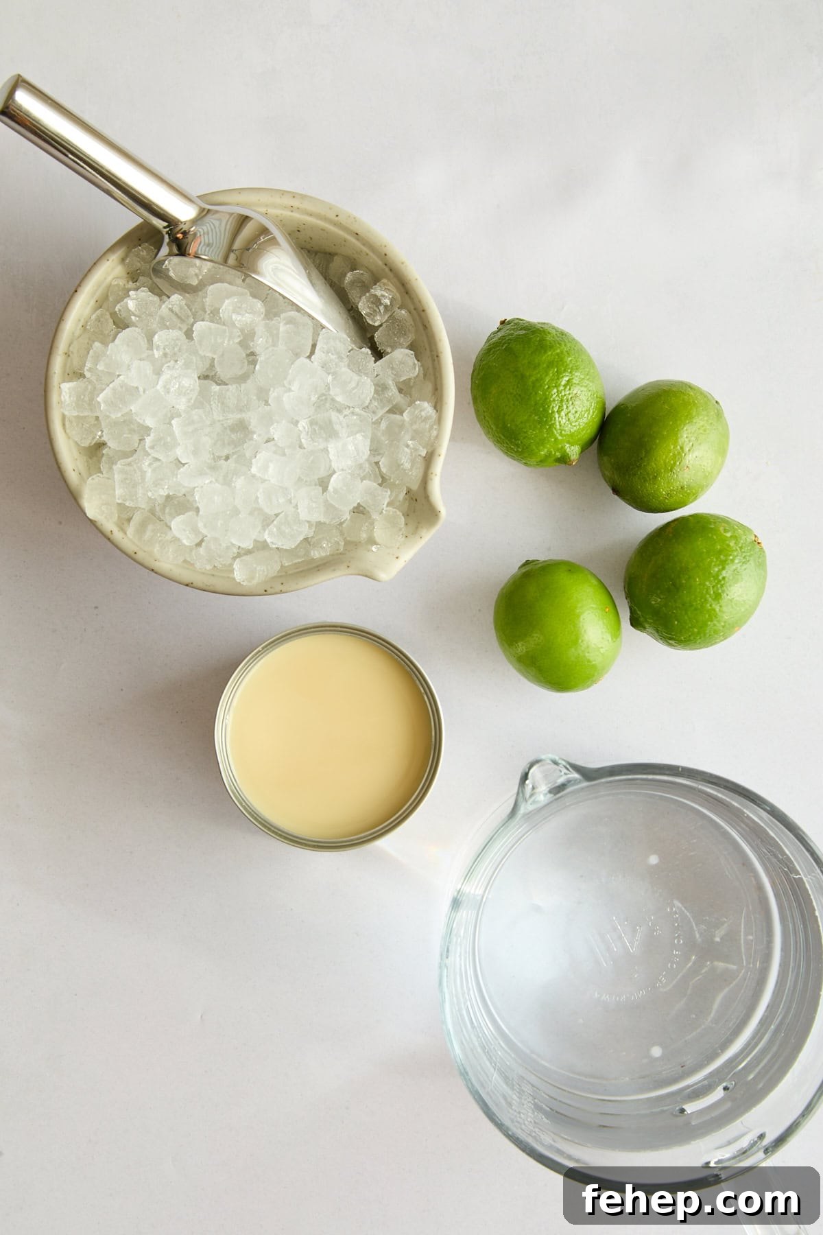 A top-down view of a bowl of crushed ice with a scoop, three whole limes, an open can of sweetened condensed milk, and a glass pitcher of water on a white surface.