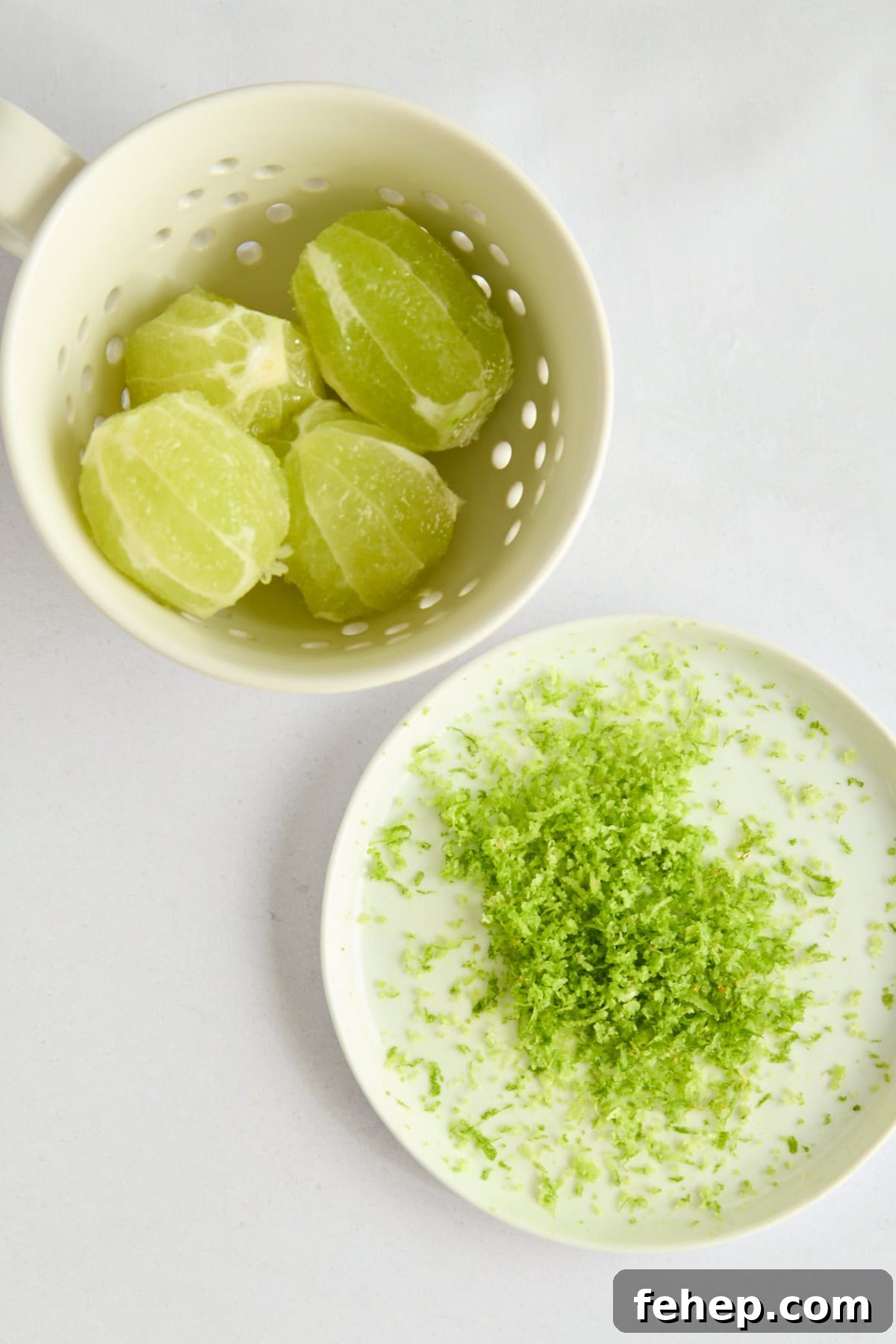 A mug with peeled limes sits above a small plate holding a pile of fresh lime zest, all on a light surface.