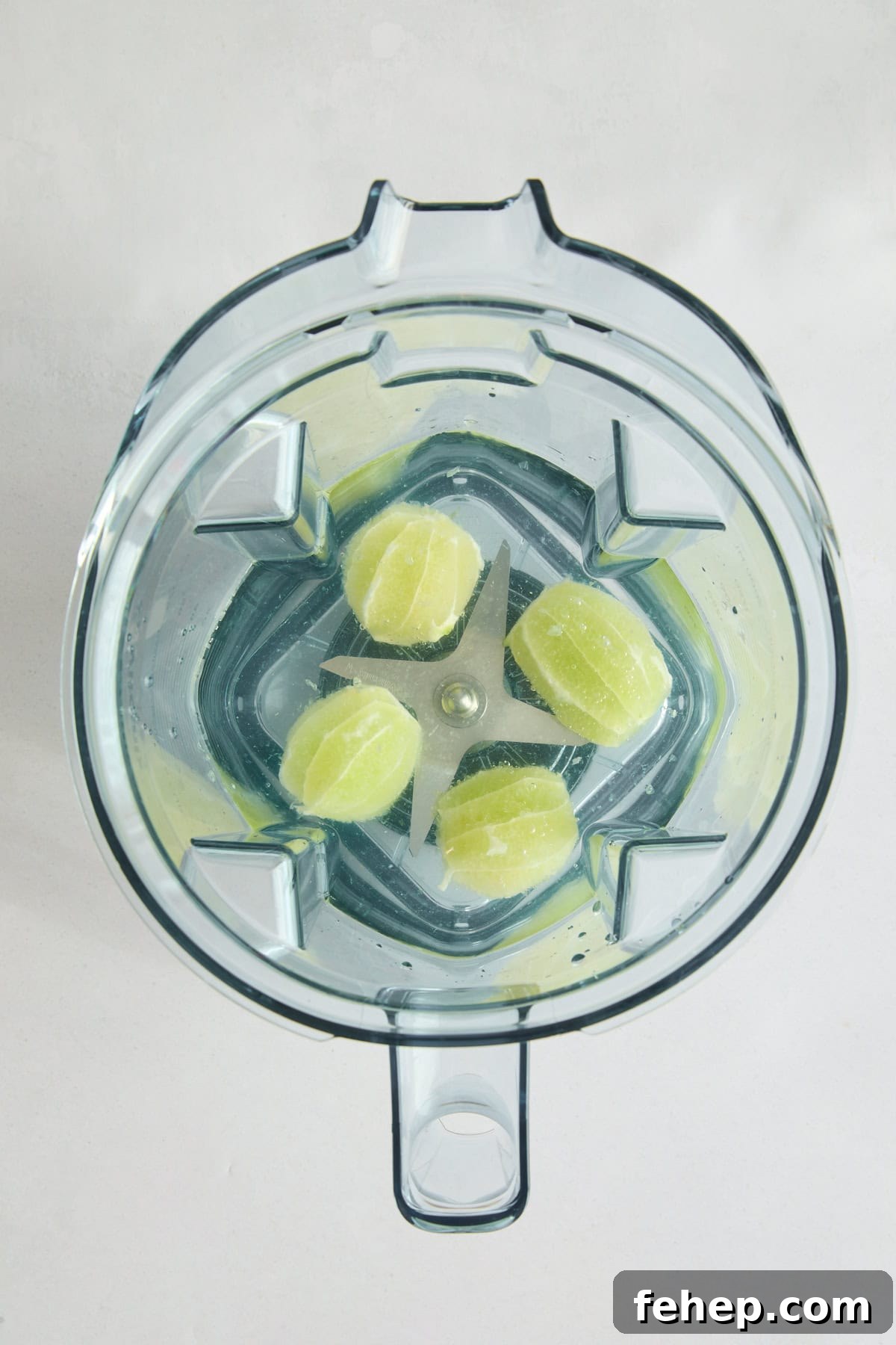 Top view of a blender with four peeled lime halves and water inside, placed on a light-colored surface.