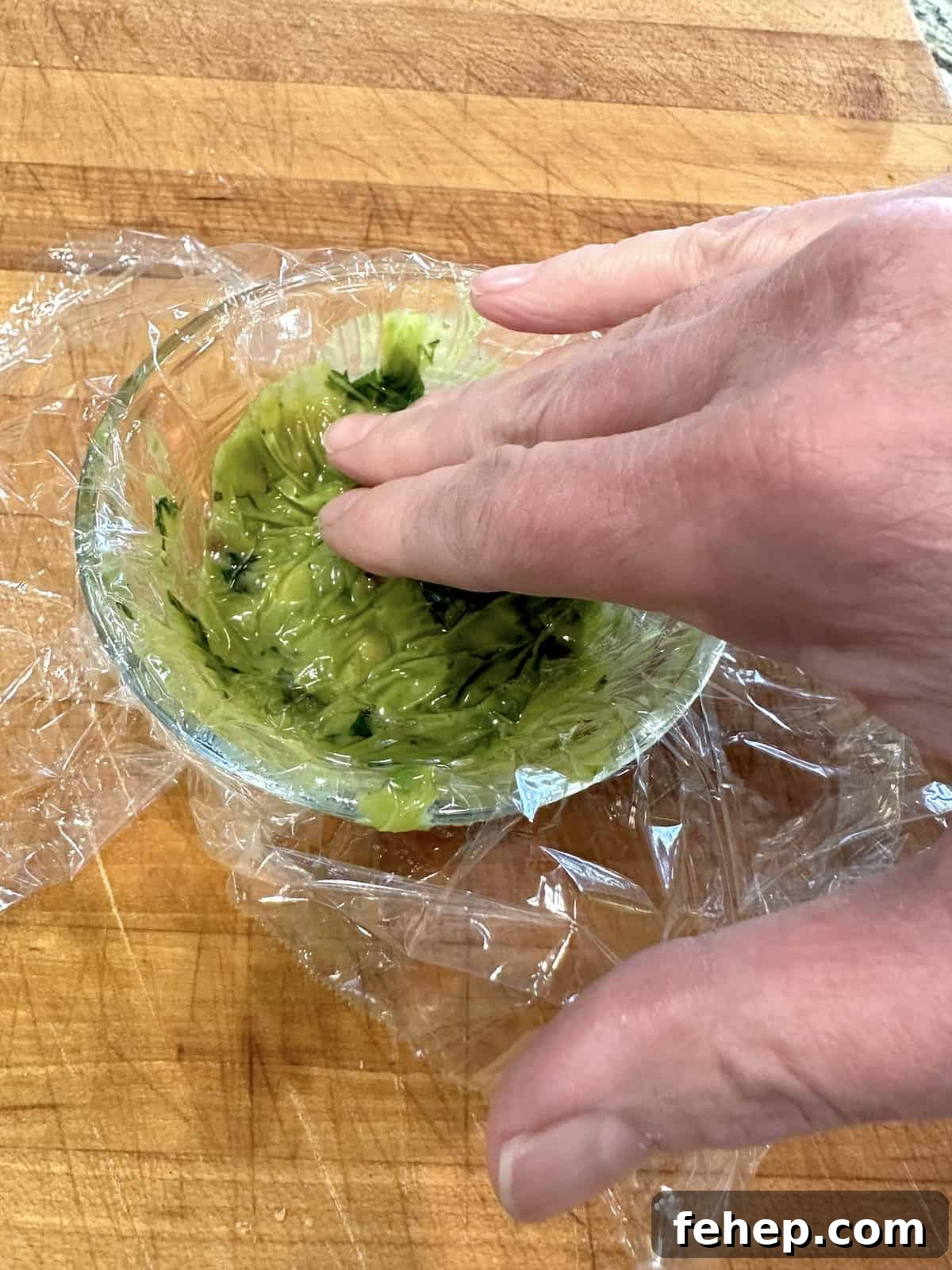 A hand pushing plastic wrap on the surface of guacamole in a bowl.