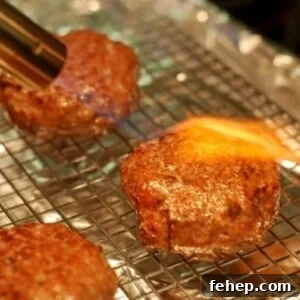 Hamburger patties made out of lamb are sitting on a wire rack. One patty has a kitchen blow torch blowing fire on it.