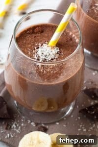 A glass of Chocolate Peanut Butter Banana Smoothie with a yellow-and-white striped straw, topped with shredded coconut and surrounded by banana slices and chocolate pieces.