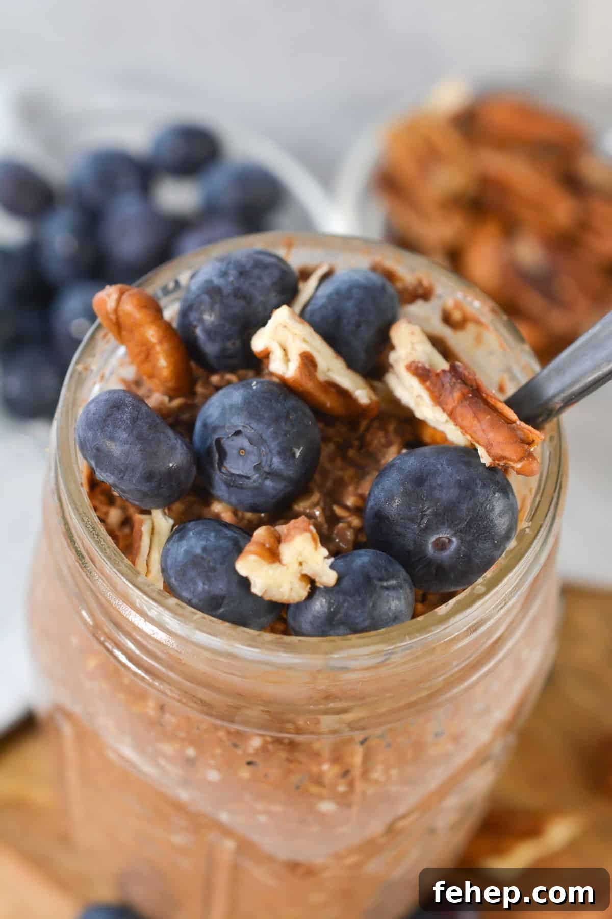 A mason jar filled with tiramisu overnight oats, topped with fresh blueberries and pecan pieces, with a spoon inside.