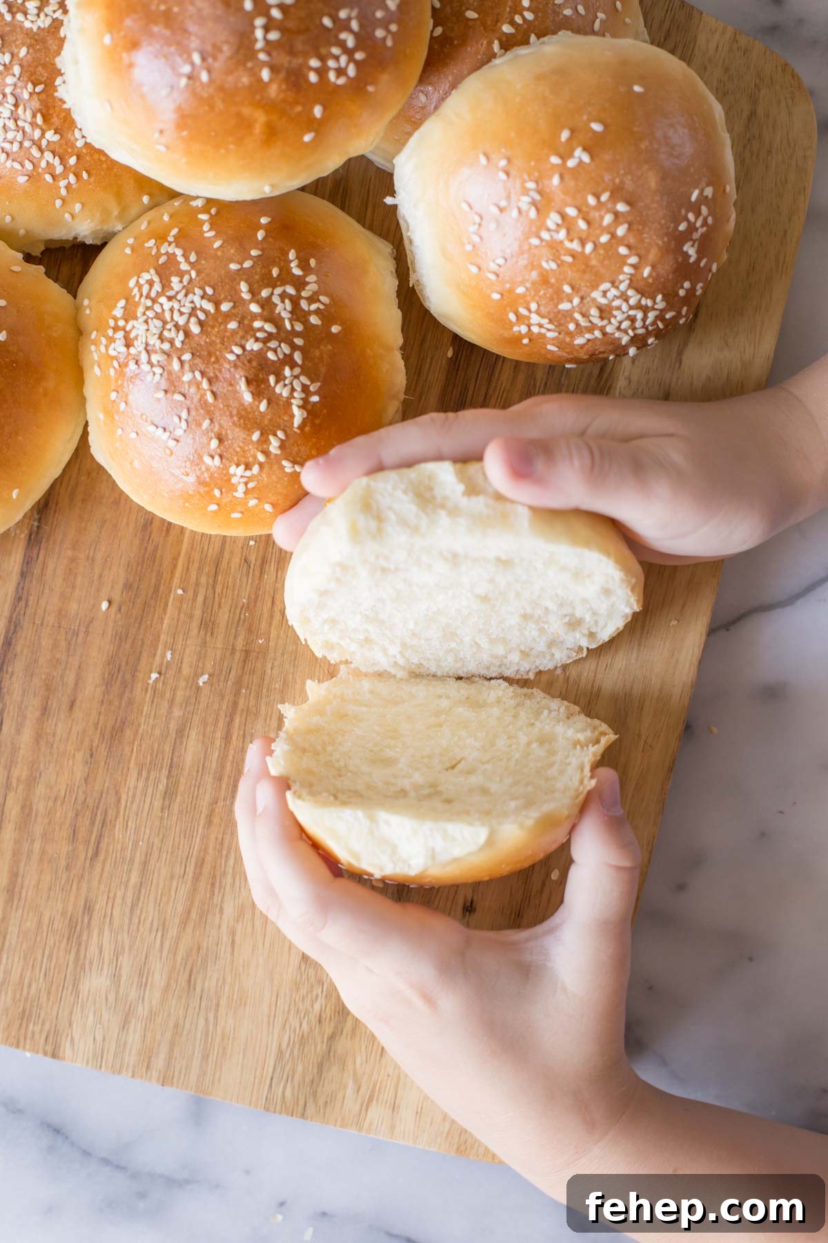 Homemade Sourdough Hamburger Buns for Tender, Tangy Burgers 5 Two hands separating a cut Sourdough Hamburger Bun on a wooden cutting board with more buns next to it.