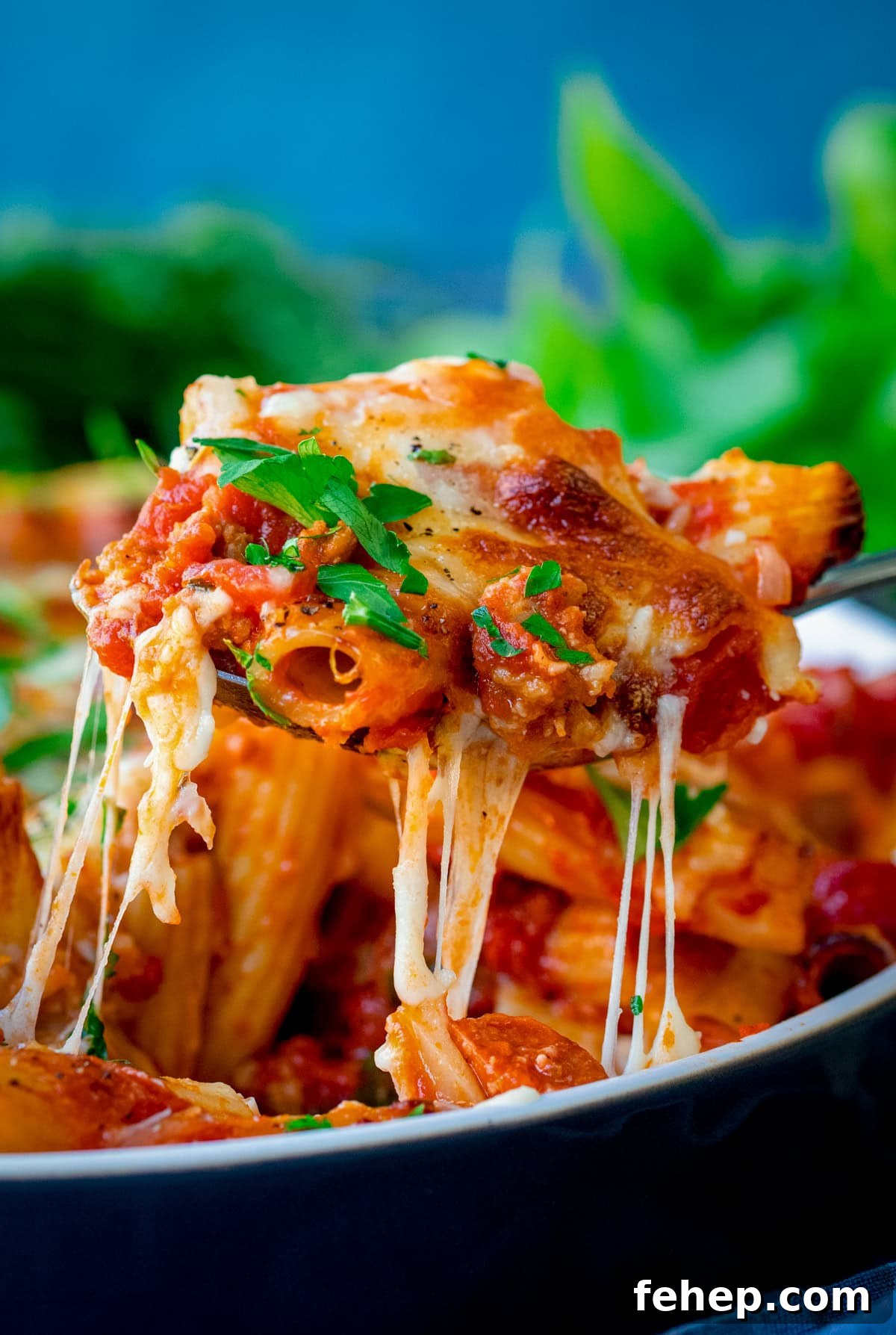 A serving spoon of Sausage Pasta Bake with tomato sauce, melted cheese, and herbs is being lifted from a dish, with gooey cheese stretching from the dish.
