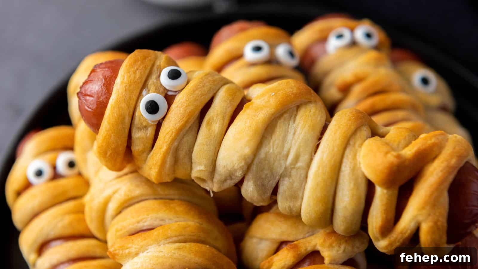 11 Spooky Halloween Treats to Liven Up Any October Night 6 Hot dogs wrapped in dough to resemble mummies, complete with candy eyes, arranged on a black plate.