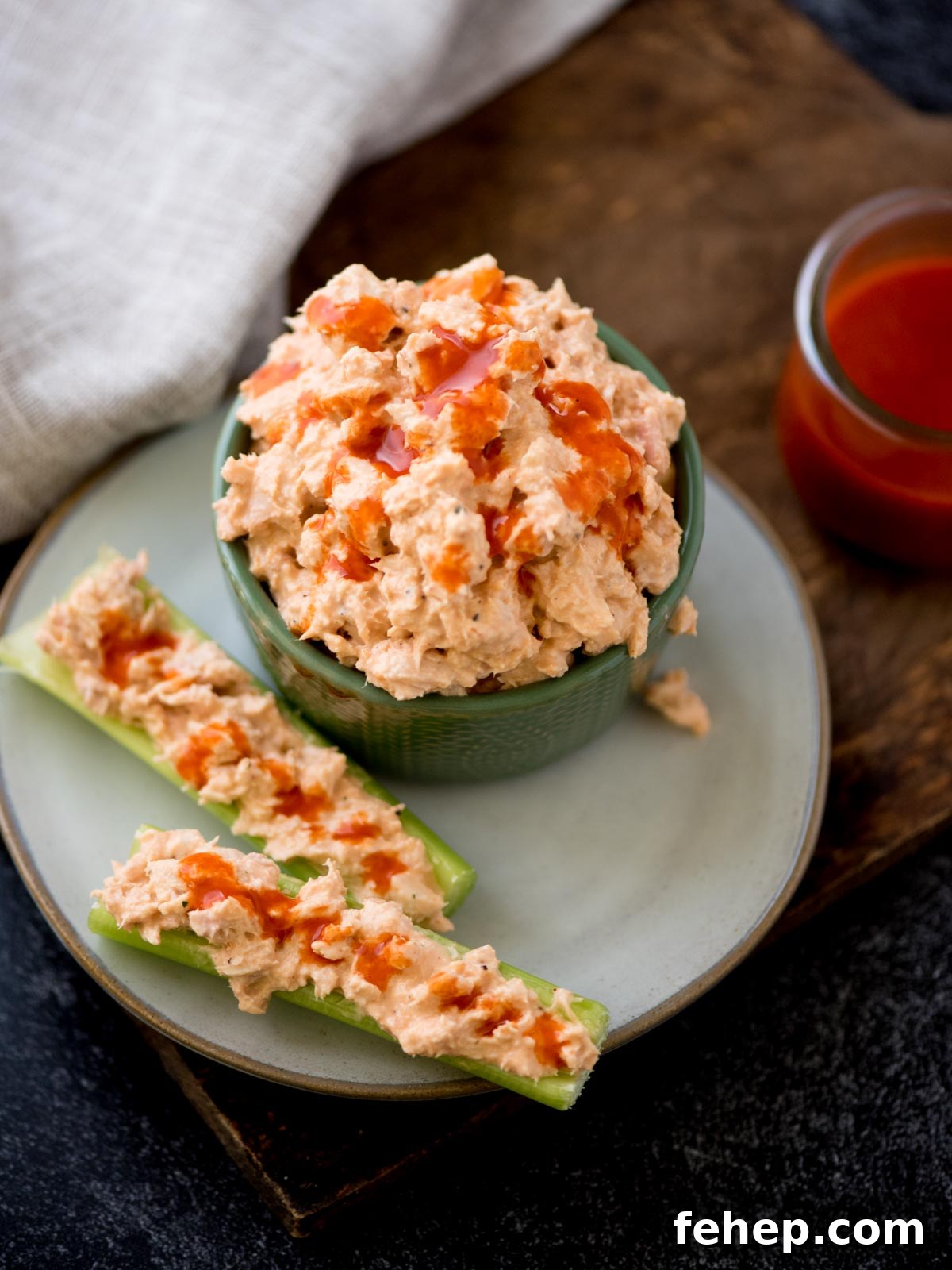 A bowl of spicy buffalo tuna salad on a plate, topped with hot sauce. Next to it are two celery sticks filled with the same dip, drizzled with hot sauce. A cloth and a small jar of spicy buffalo sauce is in the background.