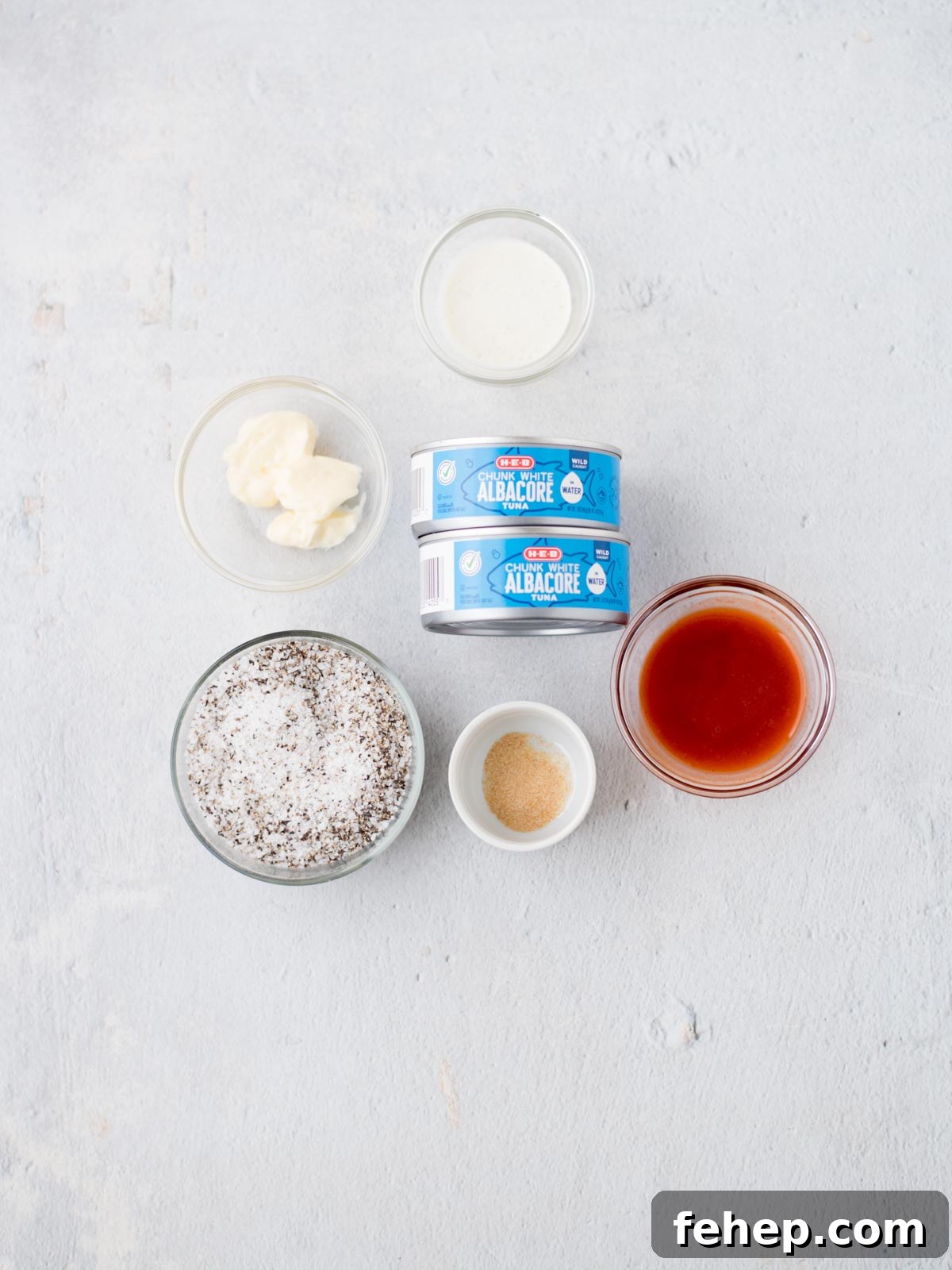 Ingredients arranged on a light surface: two cans labeled Albacore, a bowl of white sauce, a bowl of spicy buffalo sauce, a bowl of salt and pepper, and two small bowls containing liquid and powdered ingredients. Perfect for crafting a bold tuna salad with an unexpected kick.