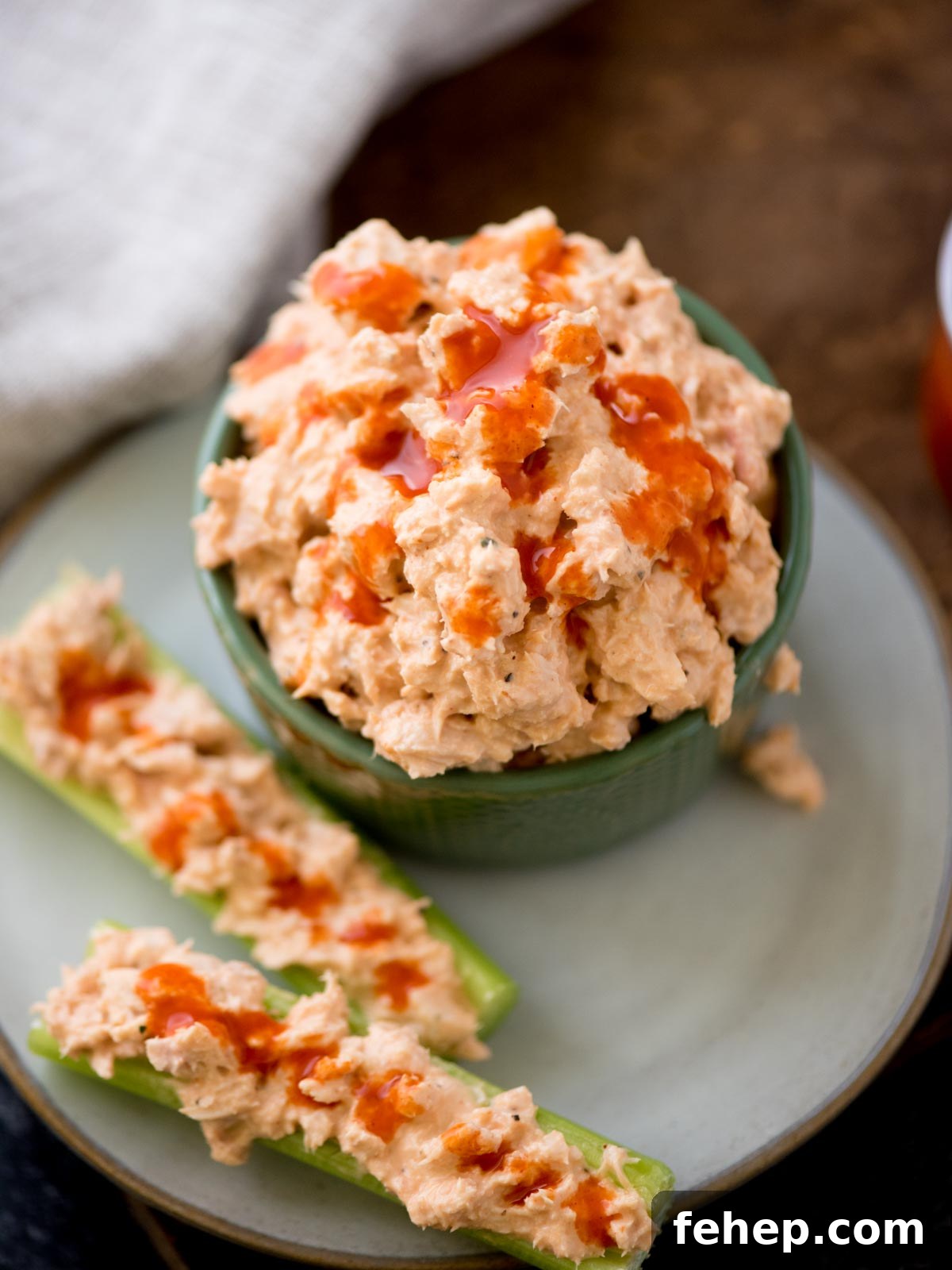 A green bowl brimming with spicy buffalo tuna dip, topped with hot sauce. On a light blue plate beside it, two celery sticks hold the same creamy mixture, drizzled with hot sauce. A light cloth graces the background.