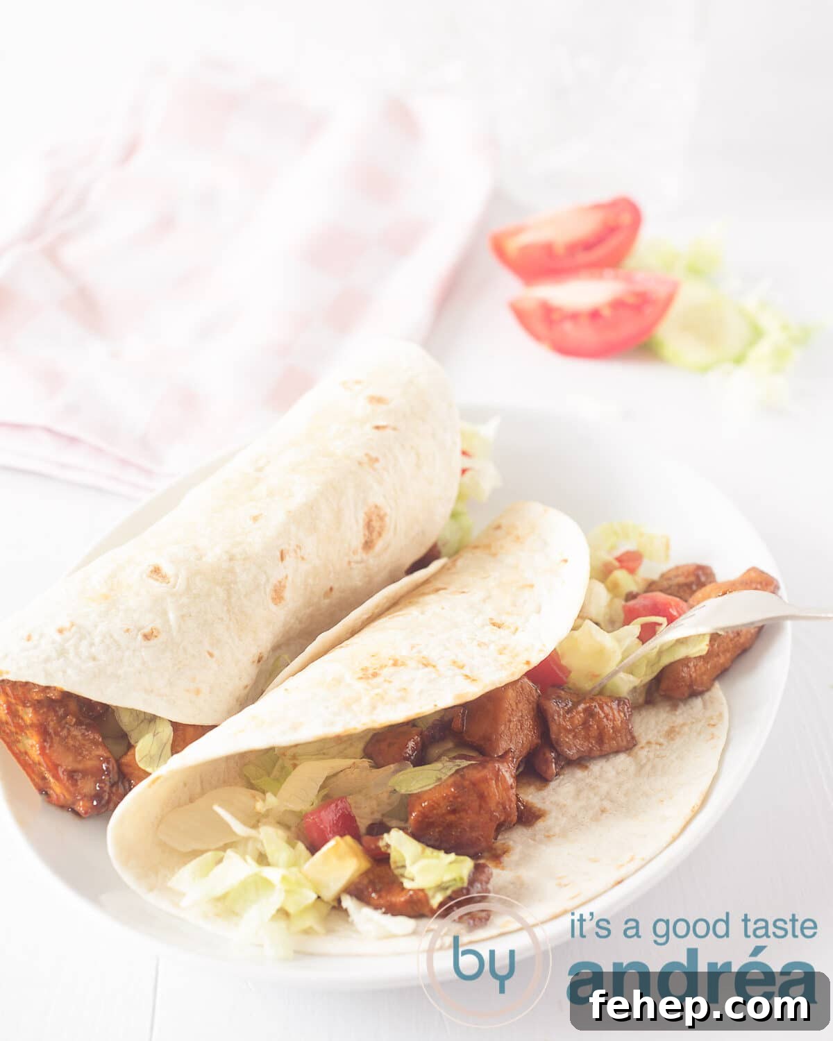Soy Chicken Wrap Recipe: Flavorful Plant-Based Lunch Ideas 2 An elevation photo with a white board. Two wraps on it. Filled with chicken, tomato, lettuce. In the background tomato wedges, lettuce and a pink white checkered tea towel