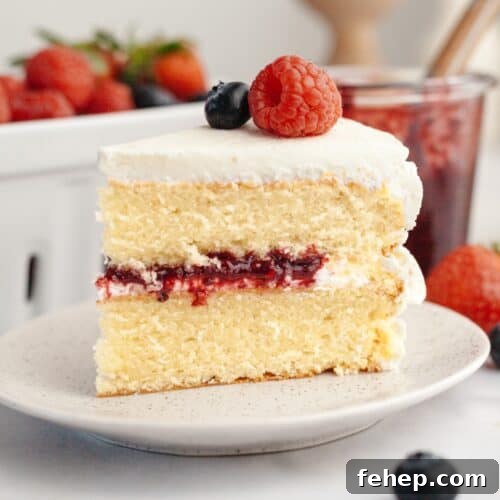 A slice of a berry vanilla layer cake with white frosting, berry jam filling, and topped with a raspberry and blueberry.