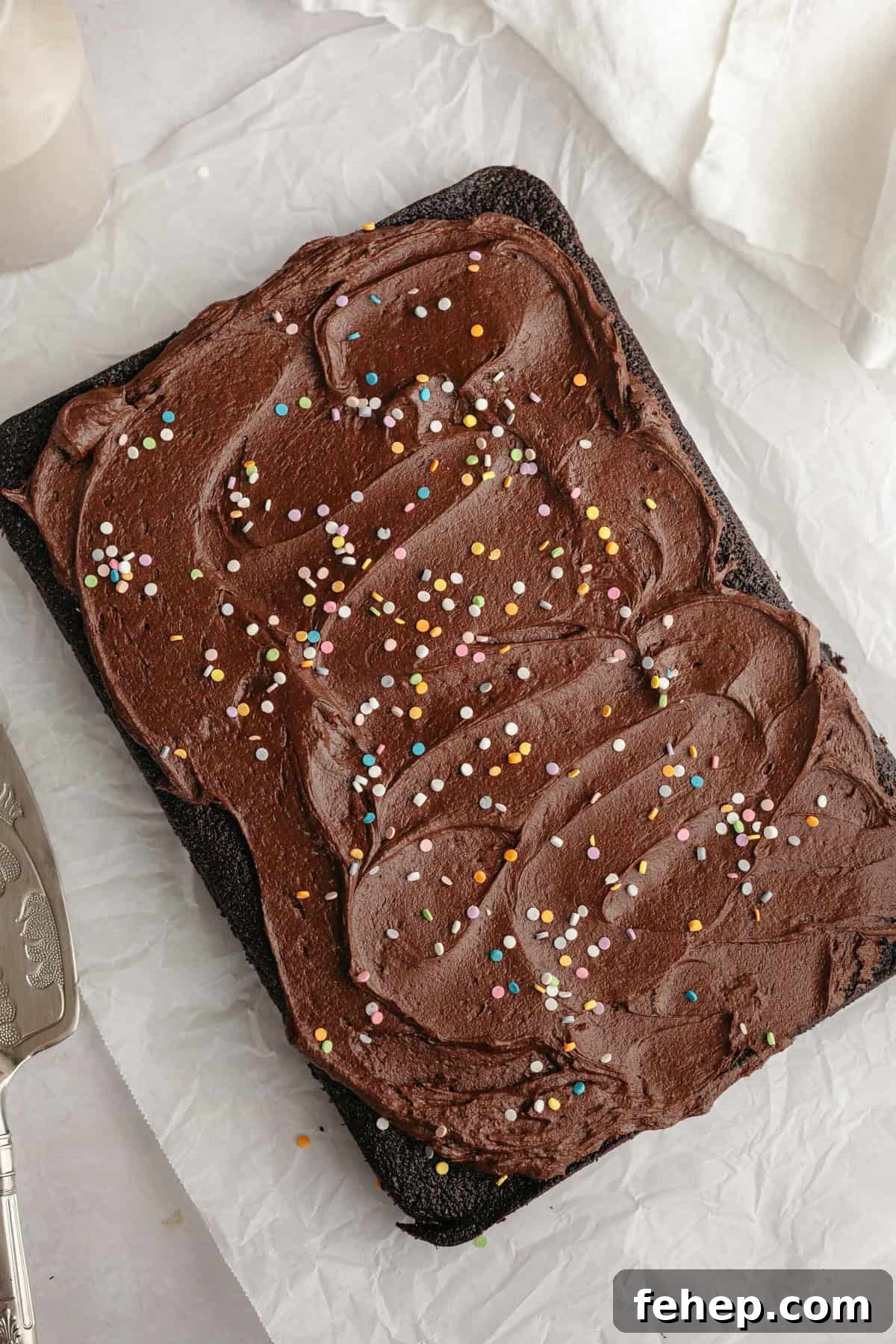 Chocolate sheet cake with chocolate frosting and rainbow sprinkles on parchment paper.