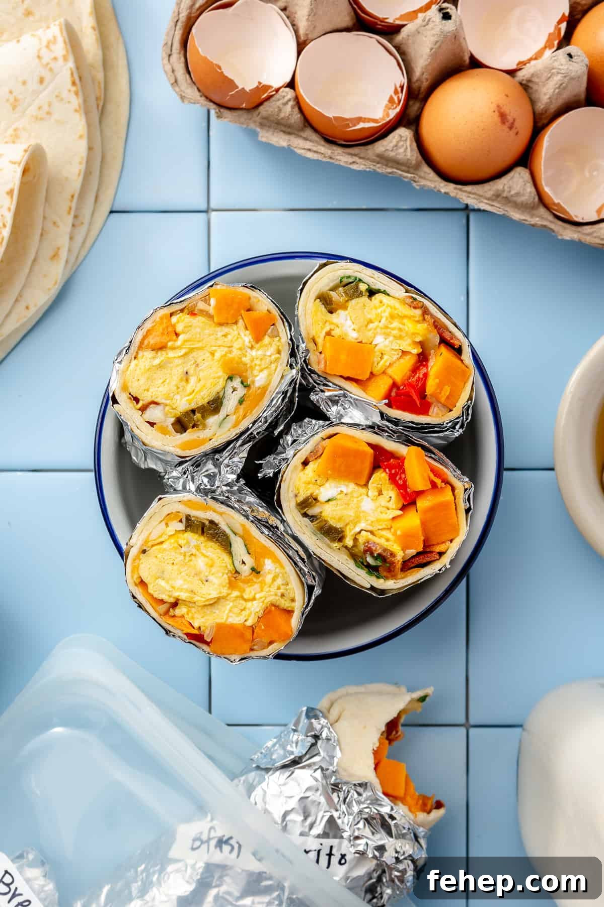 Make-Ahead Breakfast Burritos for Busy Mornings 2 Burritos cut in half and facing up on a plate in the center of photo with cracked eggs in corner and another burrito at the bottom. All on blue tile.