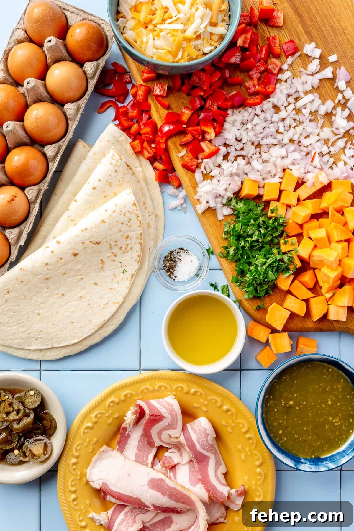 Make-Ahead Breakfast Burritos for Busy Mornings 3 Breakfast burrito ingredients. Whole eggs, tortillas, jalapeños, shredded cheese, diced red bell pepper, diced shallot, diced sweet potatoes, chopped herbs, bowl of salsa verde, and plate with uncooked bacon strips on blue tile.