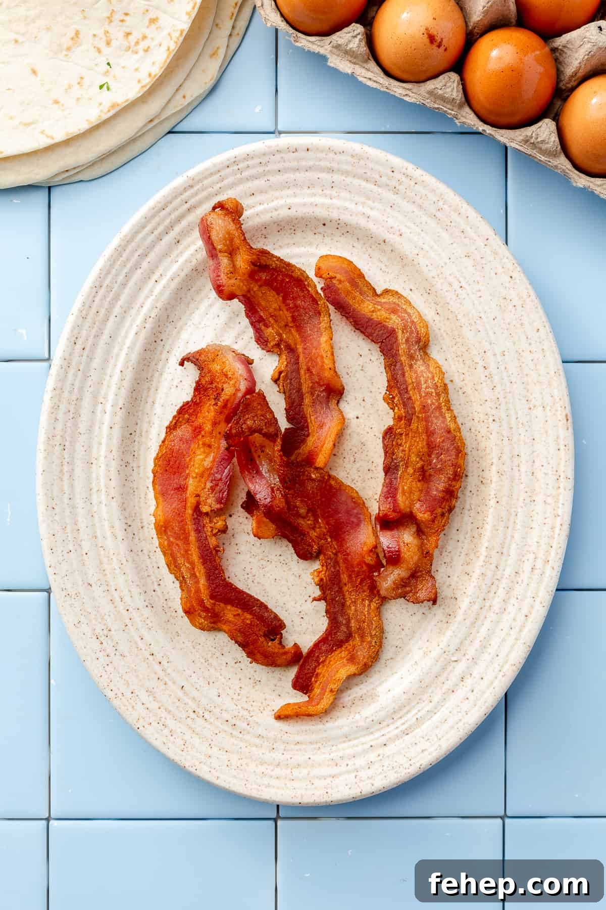 Make-Ahead Breakfast Burritos for Busy Mornings 5 Oval cream plate filled with 4 pieces of crisped bacon on blue tile backdrop.