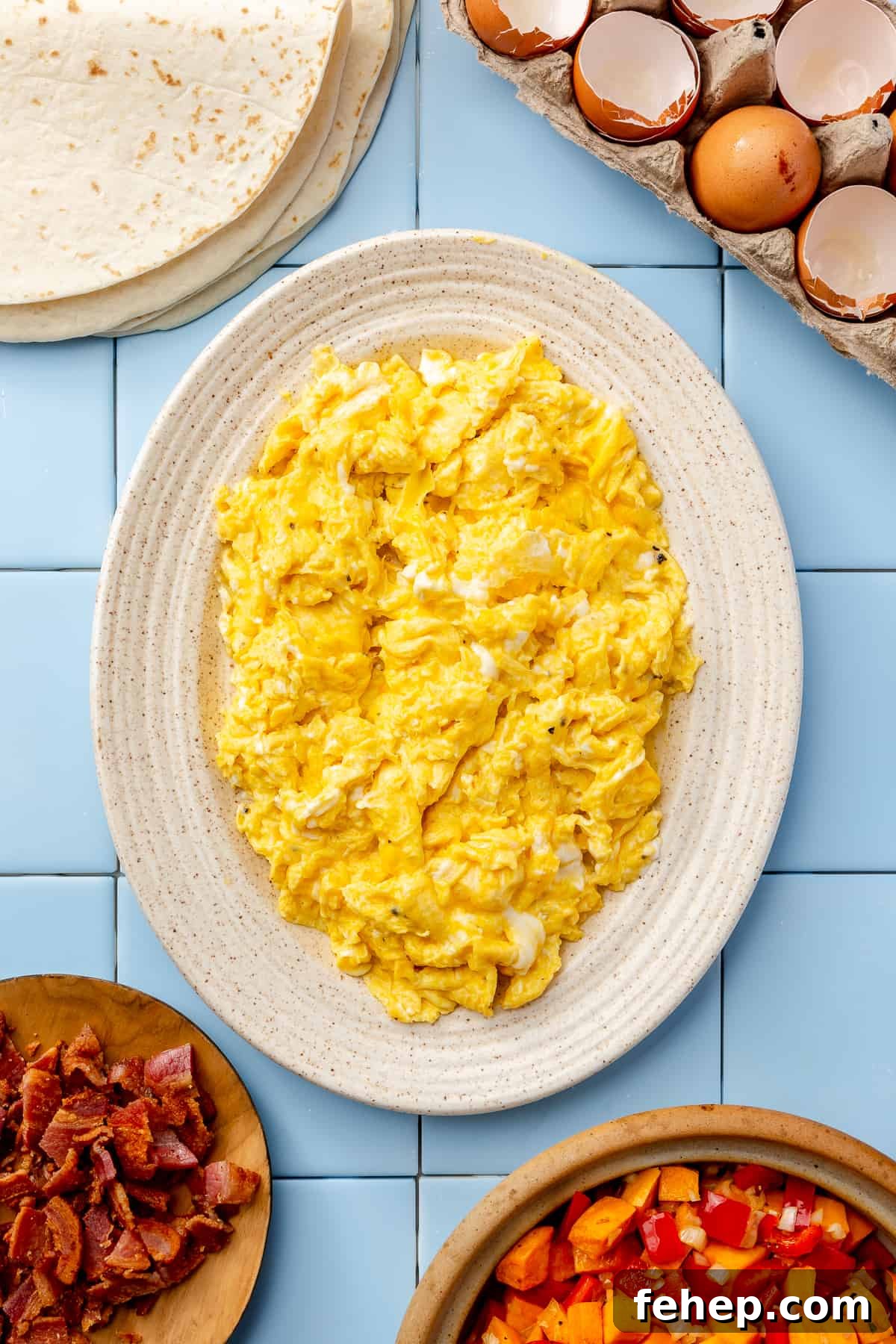 Make-Ahead Breakfast Burritos for Busy Mornings 6 Oval cream plate filled with scrambled eggs. Cracked eggs in top corner and bacon in other corner.