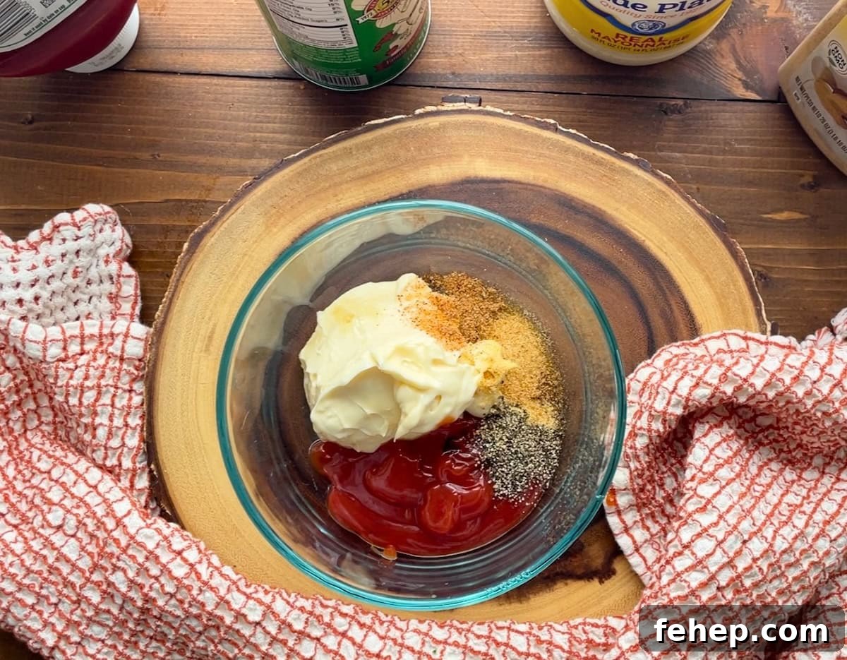 Copycat Raising Cane’s Sauce Recipe: Homemade Cane’s Dipping Sauce 5 A glass bowl containing mayonnaise, ketchup, spices, and seasonings, placed on a wooden surface with a kitchen towel beside it.