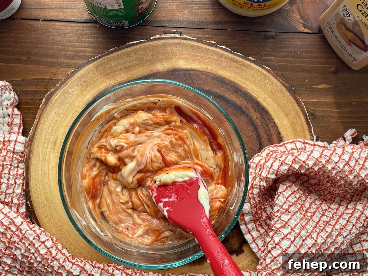 Copycat Raising Cane’s Sauce Recipe: Homemade Cane’s Dipping Sauce 6 A bowl of creamy Cane's Sauce being mixed with a spatula, surrounded by kitchen towels and condiments on a wooden table.