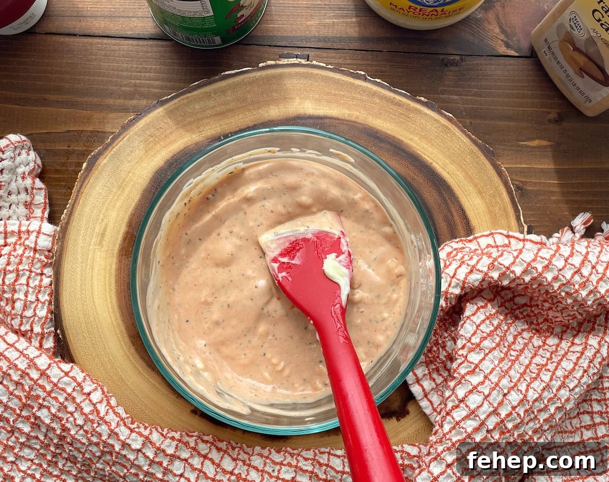 Copycat Raising Cane’s Sauce Recipe: Homemade Cane’s Dipping Sauce 7 A glass bowl filled with a creamy, pink sauce, accompanied by a red spatula, set on a wooden cutting board. A striped kitchen towel lies nearby, with various condiment bottles in the background.