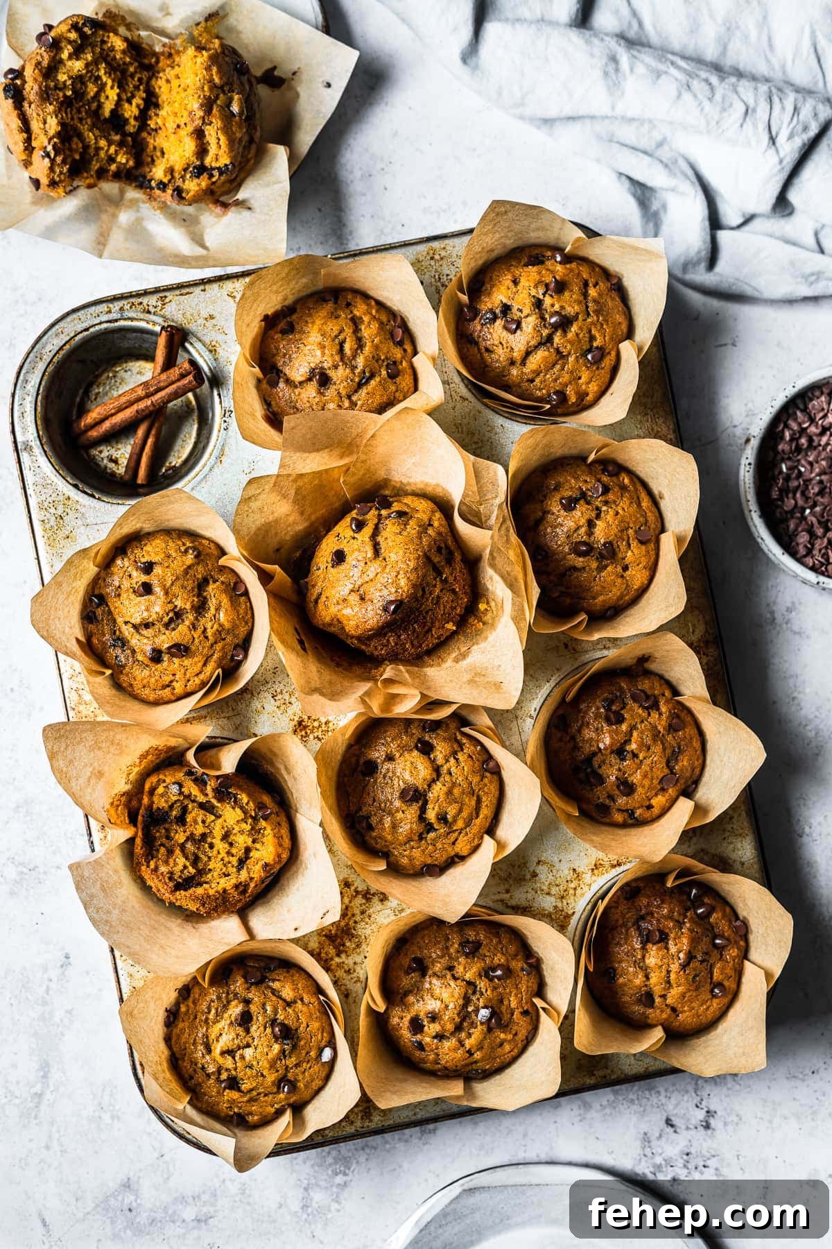 Baked pumpkin banana muffins in brown parchment liners in a muffin tin on a white stone surface. Surrounded by an opened muffin, a blue linen, a bowl of chocolate chips and a ceramic plate.