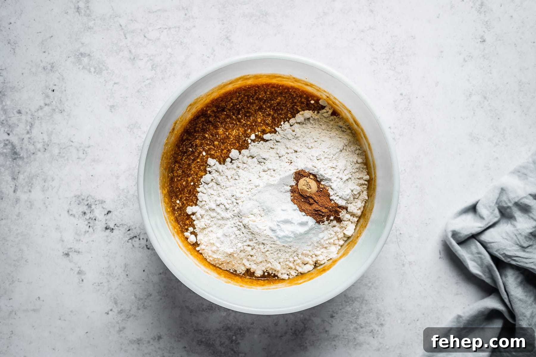 Process shot of muffin batter in a white glass mixing bowl on a textured white stone background with dry ingredients added.