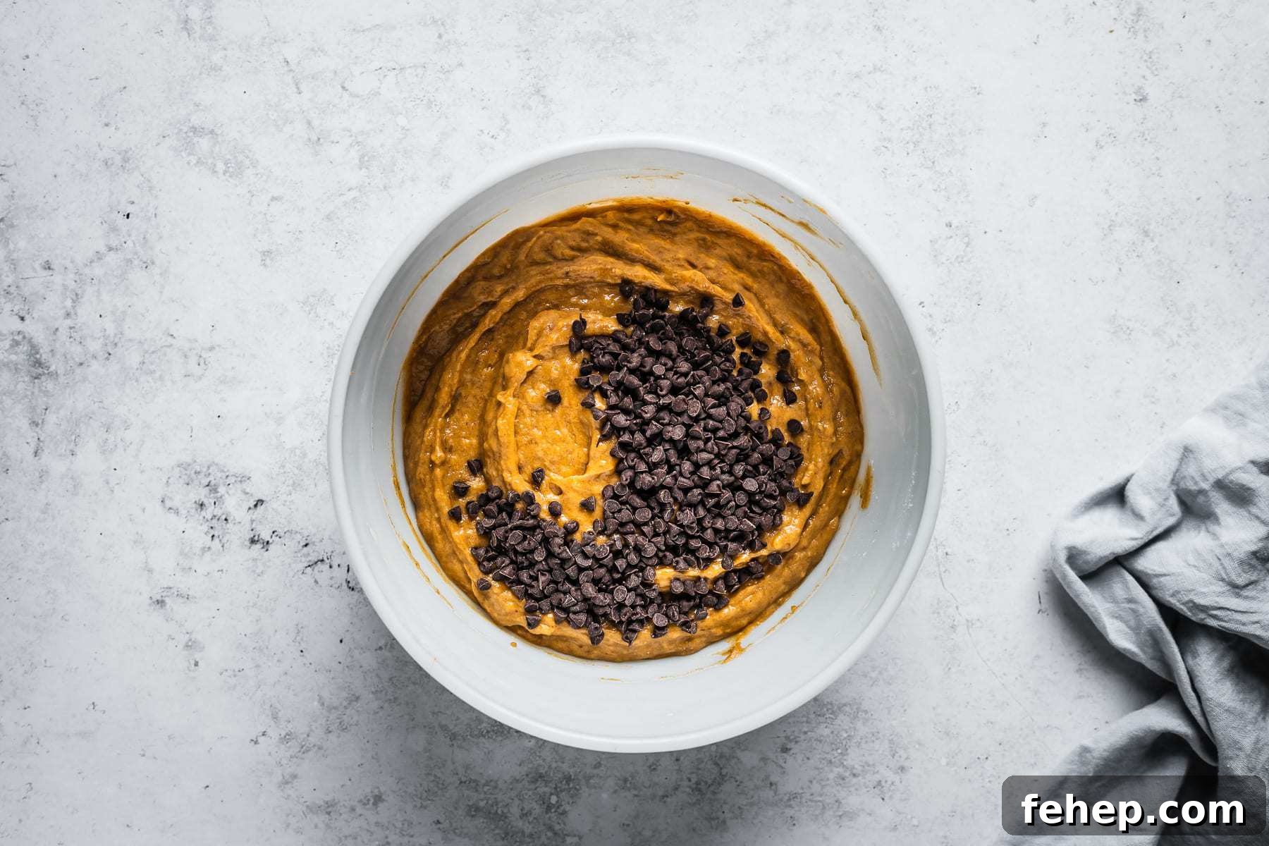 Process shot of muffin batter in a white glass mixing bowl on a textured white stone background with mini chocolate chips added.