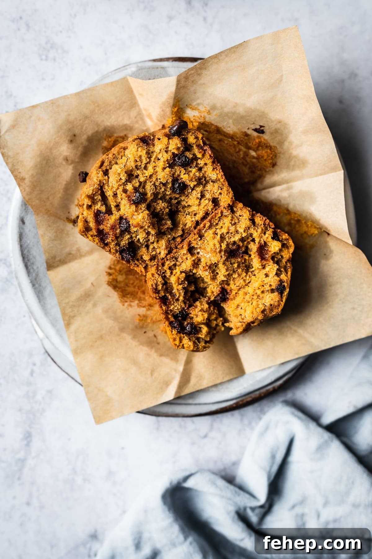 A muffin is split in half, resting face up on a brown parchment liner. It sits on a white ceramic plate on a white textured surface. A light blue linen napkin is nearby.