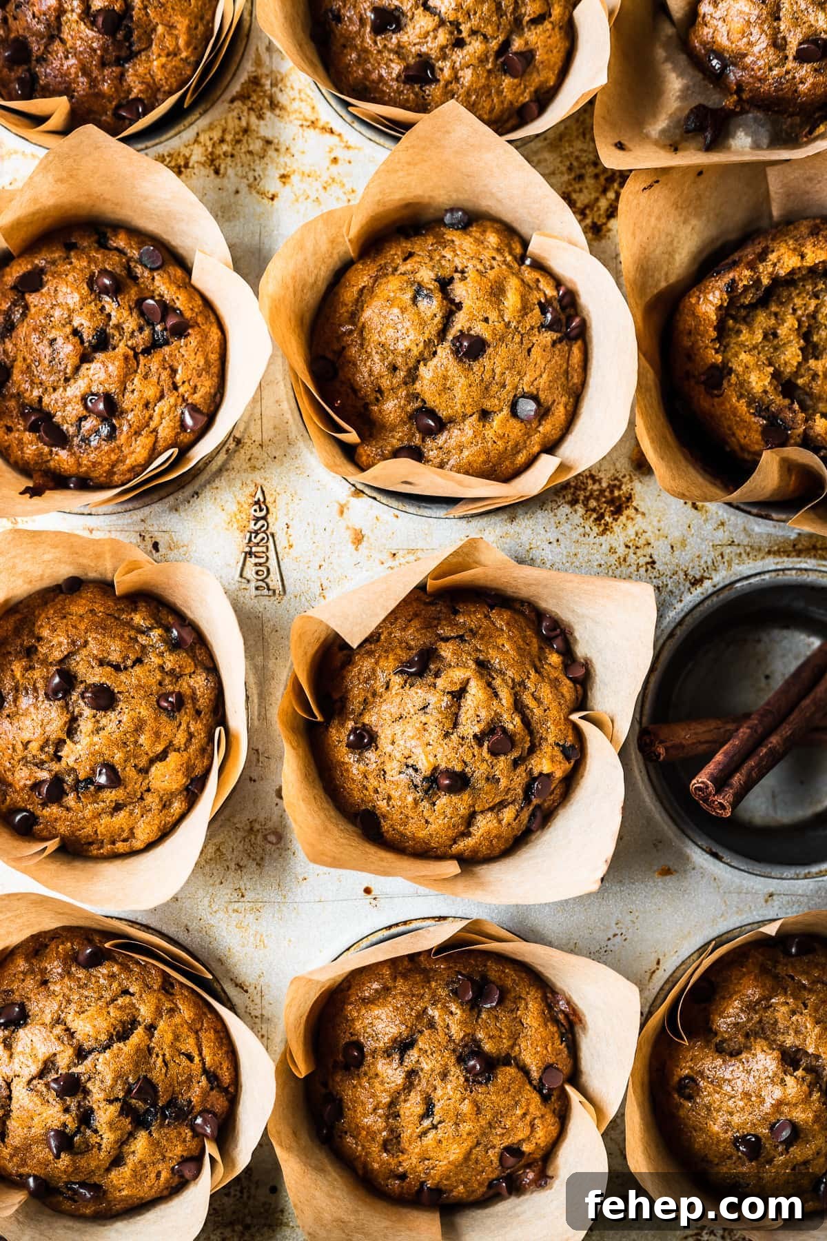 Baked muffins in brown parchment liners in a muffin tin. One cavity is empty and holds two cinnamon sticks.