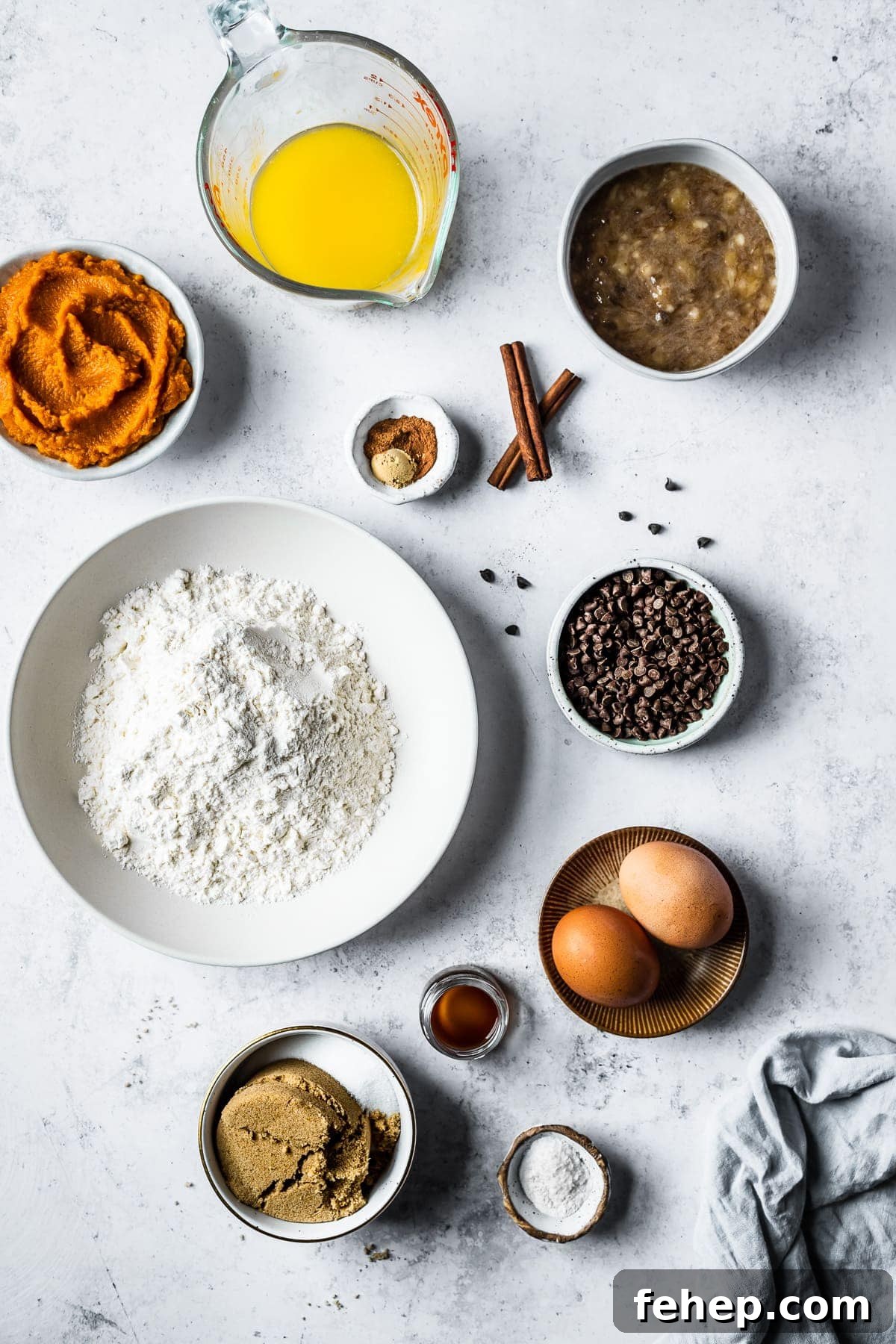 Ingredients in bowls arranged on a white textured stone surface. Ingredients include flour, pumpkin puree, melted butter, mashed bananas, eggs, sugar and spices.