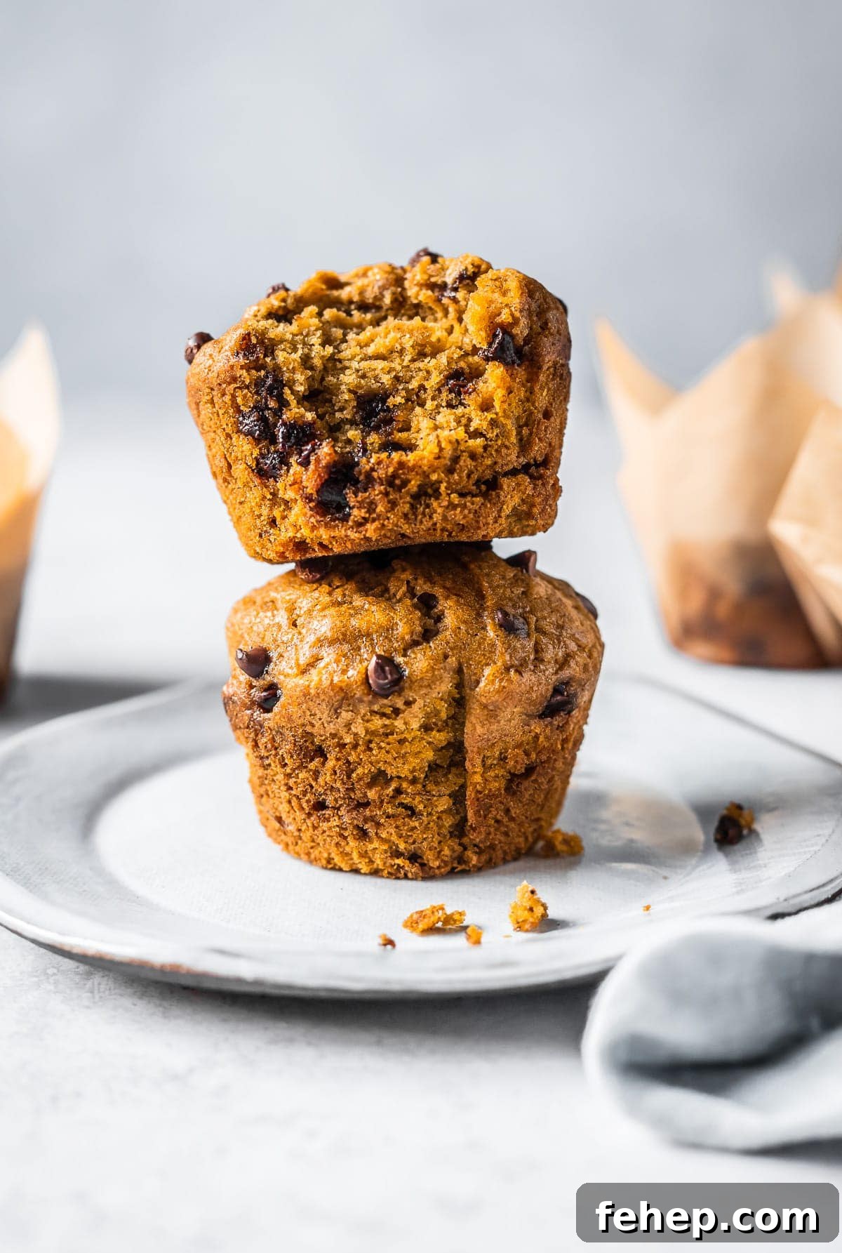 Two muffins stacked on top of each other on a white ceramic plate resting on a white stone surface. More muffins in parchment paper liners peek out from each side of the frame.