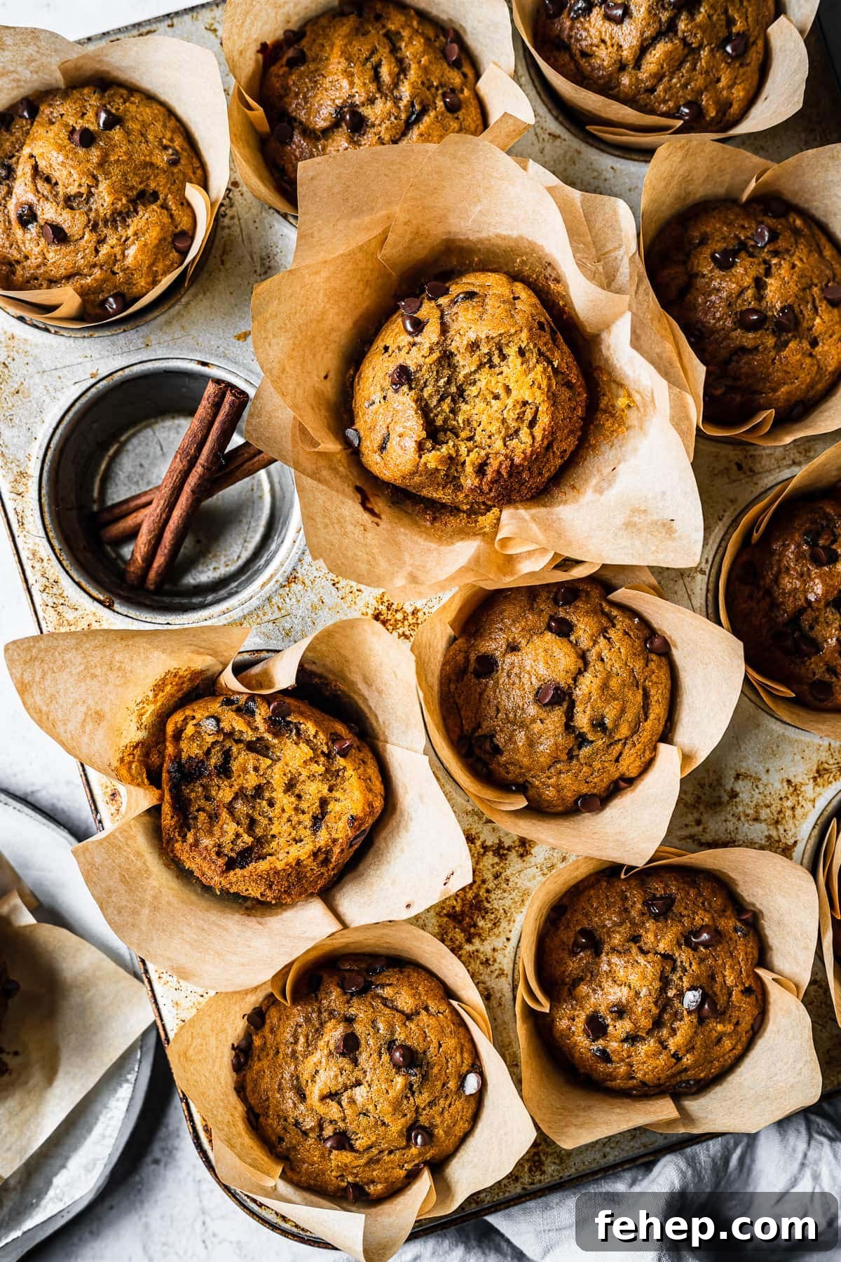Baked pumpkin banana muffins in brown parchment liners in a muffin tin. One cavity is empty and holds two cinnamon sticks. The pan is on an angle and there are bites out of two muffins, revealing the inside texture.