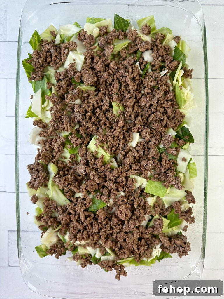 Chopped cabbage covered with cooked and crumbled ground beef in a casserole dish on a white counter top.