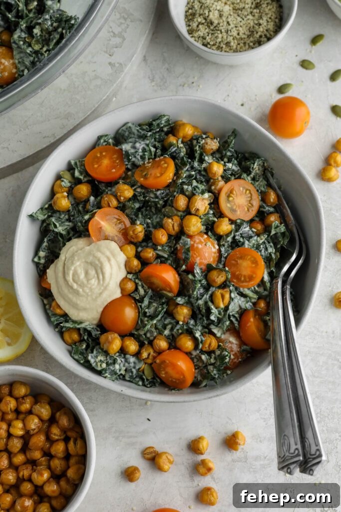Kale Caesar Salad served in a white bowl with two forks surrounded by crispy chickpeas, cherry tomatoes, hemp hearts, and pumpkin seeds