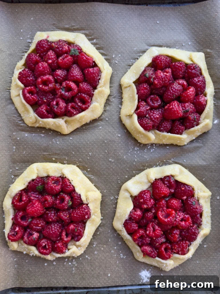 Raspberry Galette Recipe: Flaky Rustic Tarts for Summer Desserts 3 Overhead view of four unbaked raspberry filled pies on a baking sheet.