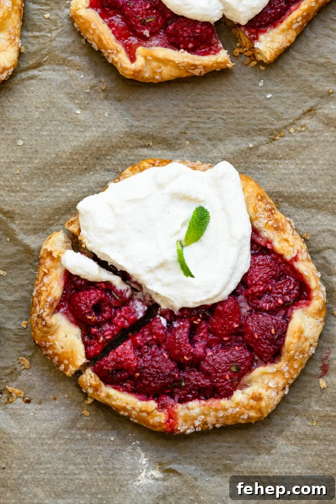 Raspberry Galette Recipe: Flaky Rustic Tarts for Summer Desserts 4 Overhead view of a hand shaped raspberry filled tart topped with cream on a baking sheet.