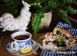 A french-toasted blackberry grilled cheese is pictured brunch style with a white rooster, a cup of tea with a slice of lemon and a spoon, a blue and white gingham tea towel and a fern in the background.