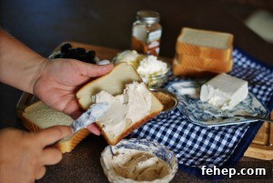 Two hands spreading fontina cheese mixed with brown sugar and cinnamon on Texas toast style bread with the ingredients for the blackberry grilled cheese are in the background on a blue and white gingham tea towel.