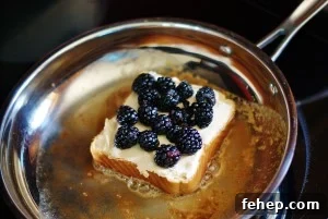 A slice of Texas toast-style bread topped with fontina cheese and blackberry french-toasting in a stainless steel skillet with browned butter.