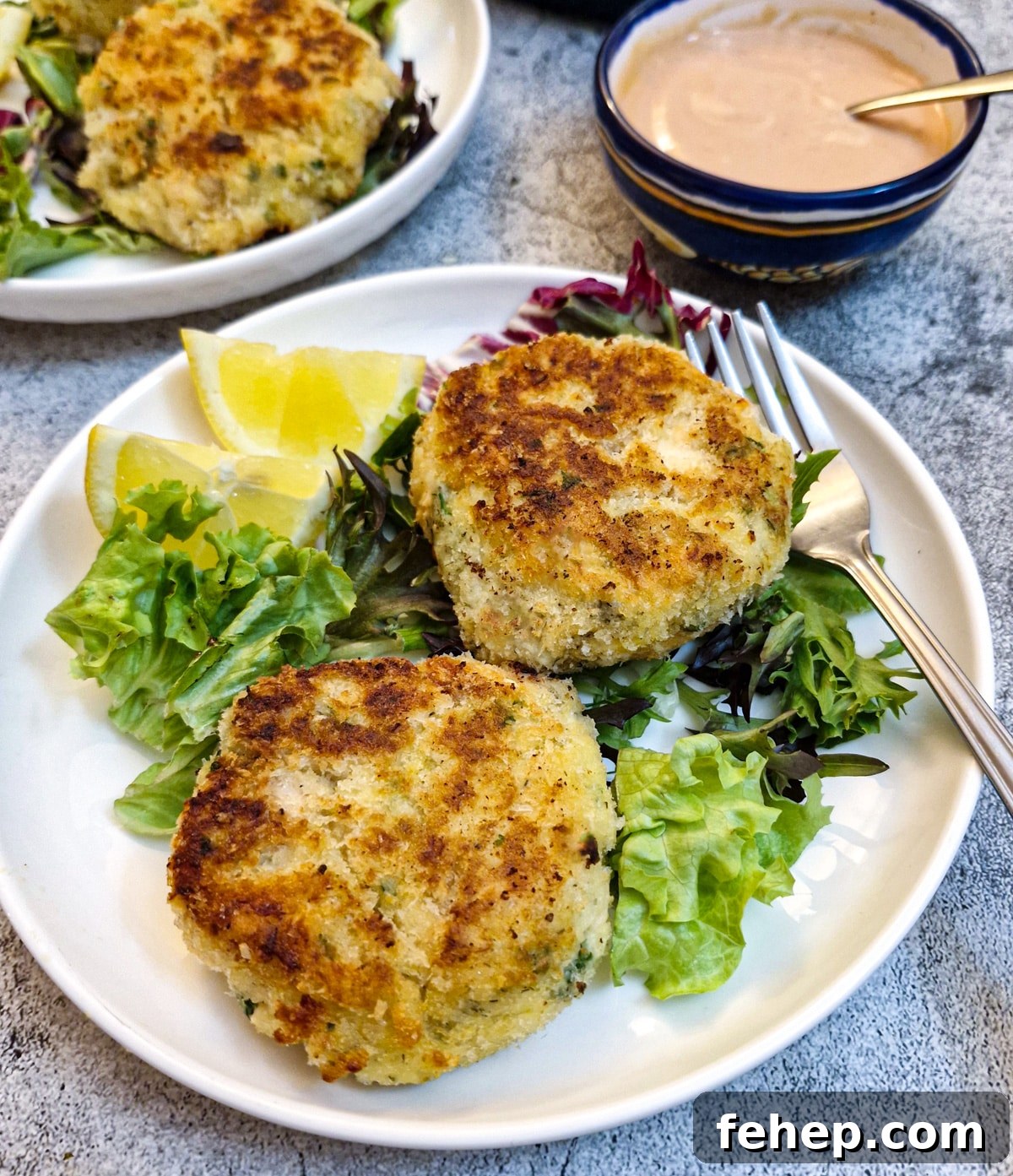 Crispy Salmon Fish Cakes with Lemon-Dill Sauce 2 Two salmon fishcakes on a white plate on a bed of lettuce with lemon slices.
