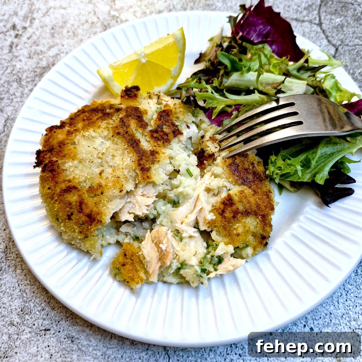 Crispy Salmon Fish Cakes with Lemon-Dill Sauce 16 A salmon fishcake which has been cut open on a white plate with lettuce and lemon wedges.