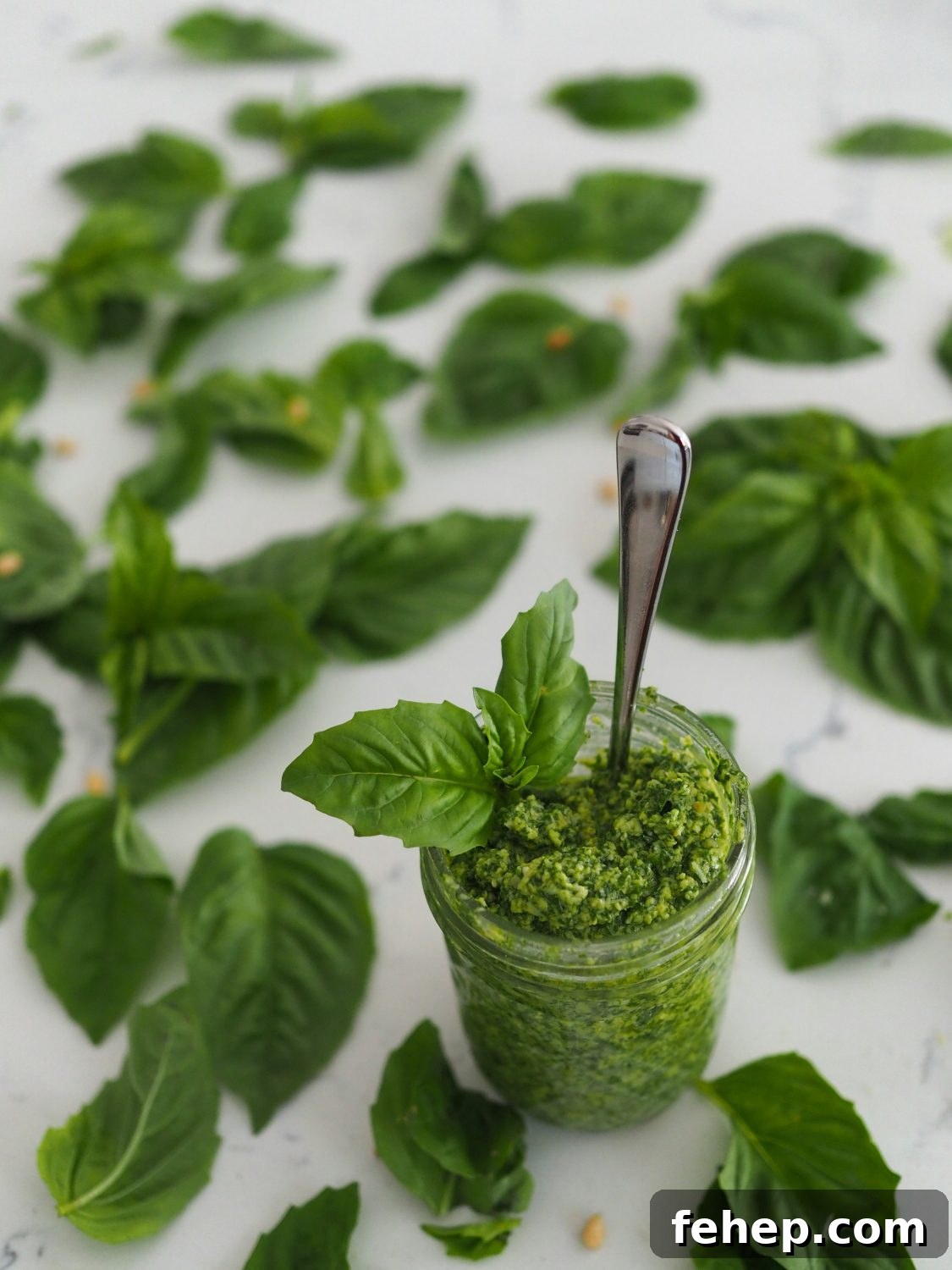 A jar of green pesto with a spoon coming out of it, surrounded by basil leaves.