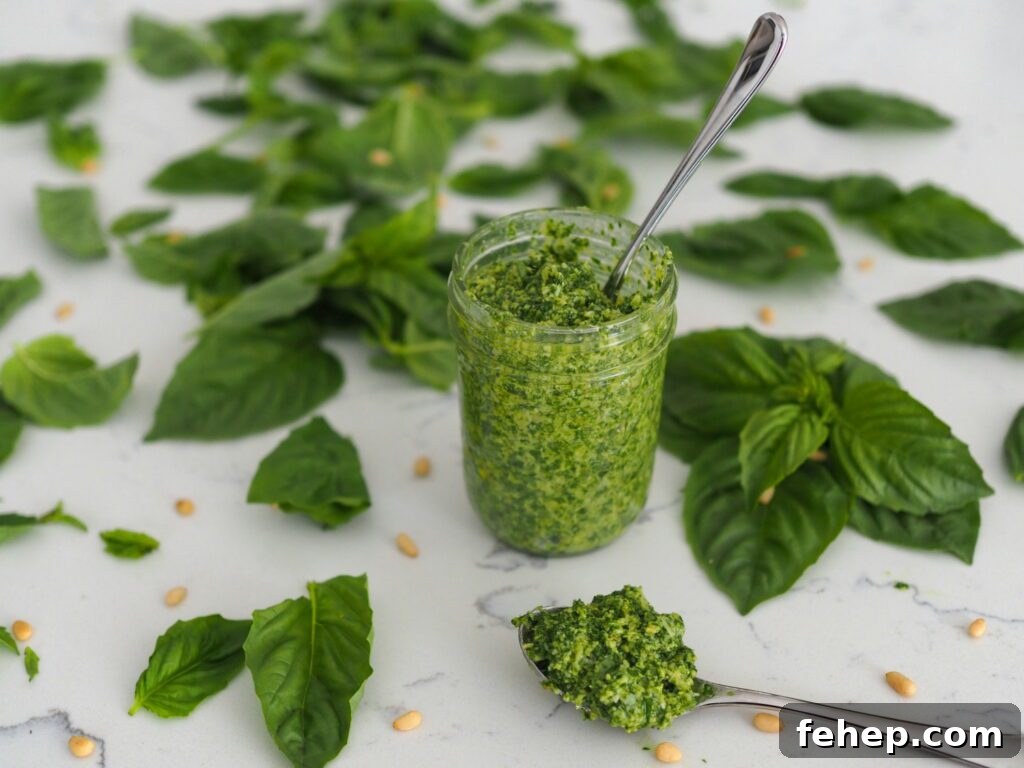 A jar of homemade pesto with a spoon sticking out, and a spoon full of pesto in front of it, with basil leaves and pine nuts around them.