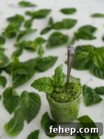 A jar of green pesto with a spoon coming out of it, surrounded by basil leaves.