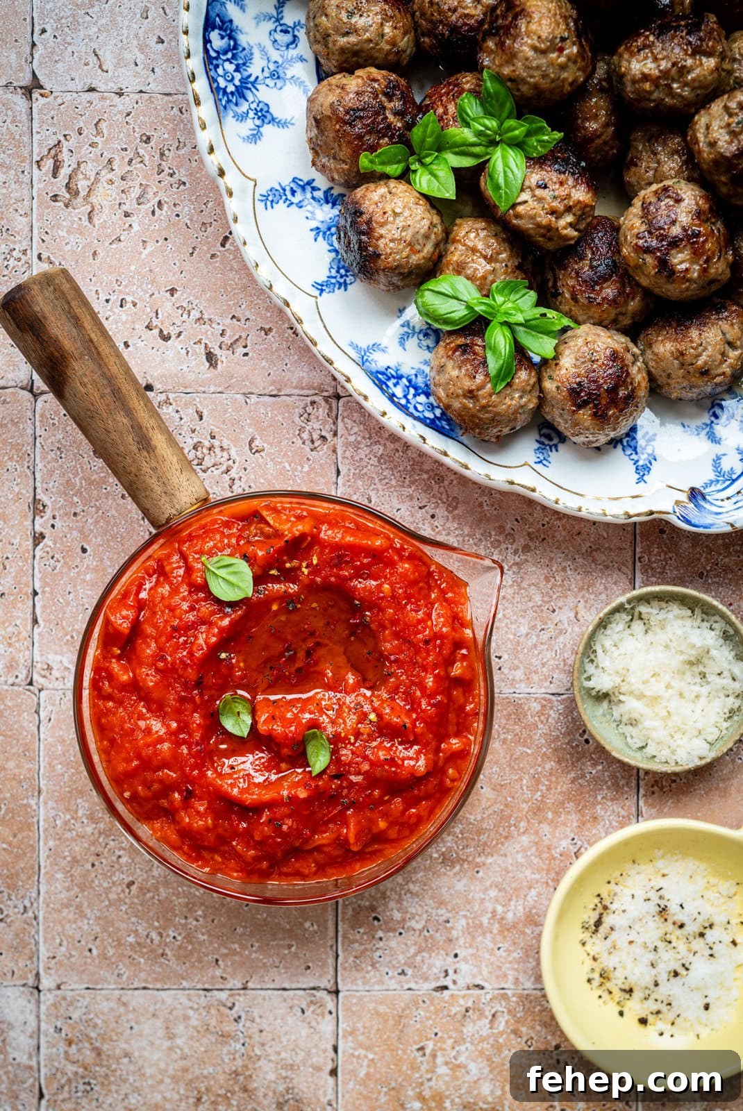 Authentic San Marzano Tomato Sauce Recipe for Rich Flavor 3 A bowl of chunky tomato sauce garnished with basil sits beside a plate of meatballs topped with fresh basil, with small bowls of grated cheese and olive oil on a tiled surface.