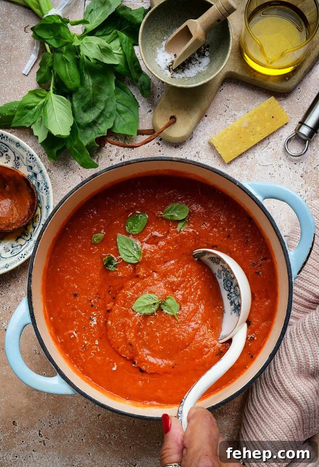 Authentic San Marzano Tomato Sauce Recipe for Rich Flavor 7 A hand ladles rich tomato soup from a blue pot, garnished with fresh basil leaves. Surrounding the pot are basil, olive oil, salt, a cheese wedge, and a small plate with more sauce.